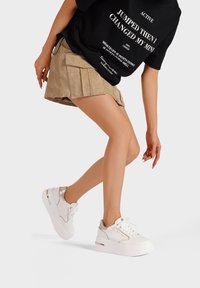 Sneaker platform bianchi con accenti metallici, tomaia in pelle liscia e suola testurizzata, abbinati a una maglietta nera e una gonna plissettata beige.