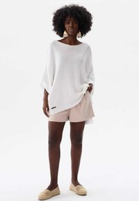Weißer, gestrickter Poncho mit lockerer Passform, der eine diagonale Linie offener Maschen und ein kleines Logoband aufweist, kombiniert mit hellbeigen Shorts.