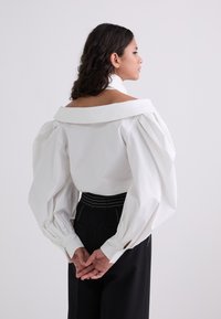 Blusa bianca a spalle scoperte con maniche voluminose, colletto strutturato e una texture liscia. Abbinata a pantaloni neri a vita alta.