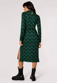 Abito verde a maniche lunghe con motivo geometrico, caratterizzato da una vita stretta, fessure laterali e un'apertura sul collo sul retro. Indossato con stivaletti neri.