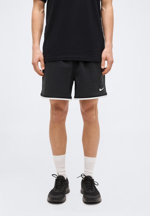 Άτομο που φοράει μαύρο αθλητικό σορτς με λευκή επένδυση και λογότυπο, μαύρο μπλουζάκι, λευκές κάλτσες και μαύρα αθλητικά παπούτσια, όρθιο σε απλό φόντο.