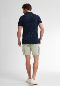 Námořnické polo tričko s malými žlutými a zelenými vzory, spárované se světle zelenými cargo kraťasy a bílými teniskami. Klasický design, krátké rukávy.