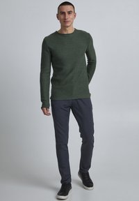 Grön stickad tröja med rund halsringning, bärs med mörkgrå slim fit byxor och svarta sneakers, mot en ljus bakgrund.
