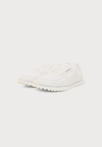 Paire de baskets basses blanches avec un design à lacets et des semelles texturées sur un fond clair uni.