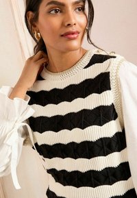 Knitted sweater in black and cream stripes, featuring a textured pattern, round neckline, and decorative buttons on the shoulder.