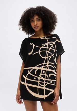 Femeie cu păr creț, purtând o rochie tunică neagră cu un model abstract de linii bej, stând pe un fundal simplu și deschis la culoare.