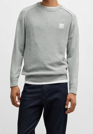 Mand iført en lysegrå tekstureret crewneck-sweater med et lille BOSS-logo, mørke jeans og en hvid skjorte under.