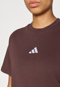 Burgundinės medvilninės marškinėliai su apvaliu iškirpimu; juos puošia baltas siuvinėtas logotipas, sudarytas iš trijų įstrižai nukreiptų juostų krūtinėje.
