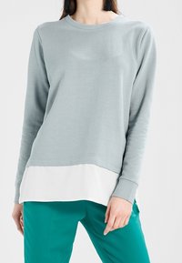Sudadera de manga larga gris claro con un dobladillo blanco en capas, hecha de una tela suave, combinada con pantalones en tono verde azulado. Diseño simple y corte relajado.