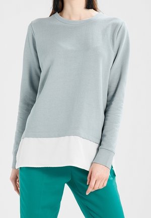 Felpa grigia chiara a maniche lunghe con orlo bianco a strati, realizzata in tessuto morbido, abbinata a pantaloni color teal. Design semplice e vestibilità casual.