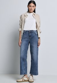 Chemise en velours côtelé beige clair, t-shirt blanc, jeans évasés bleus, et baskets crème avec des accents bruns. Fermeture à boutons simple sur la chemise.
