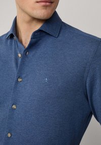 Hombre vestido con una camisa de botones azul con un pequeño logo bordado en el pecho, con el cuello abierto, mostrando parte del cuello y la barbilla.
