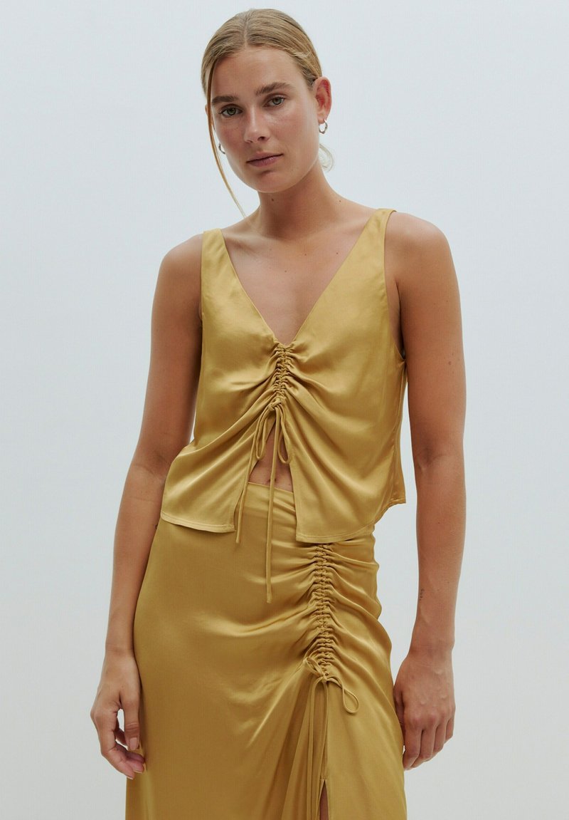 Gouden satijnen top met een diepe V-hals en geplooide details in het midden. De stof is glad en glanzend, wat de elegante silhouet benadrukt.