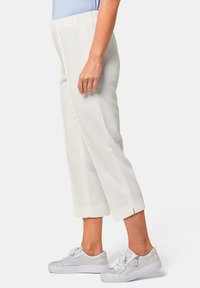 Witte cropped broeken van een textuurstof, met een elastische tailleband en split hem, gecombineerd met zilveren sneakers. Zijaanzicht dat de relaxte pasvorm toont.