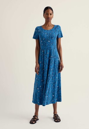 Nő kék virágmintás midi ruhában, rövid ujjú, fekete pántos szandálokkal, egyszerű bézs háttér előtt állva.