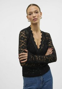 Top en dentelle noire avec un décolleté en V profond, manches longues avec des poignets évasés, motifs floraux complexes et silouhette ajustée.