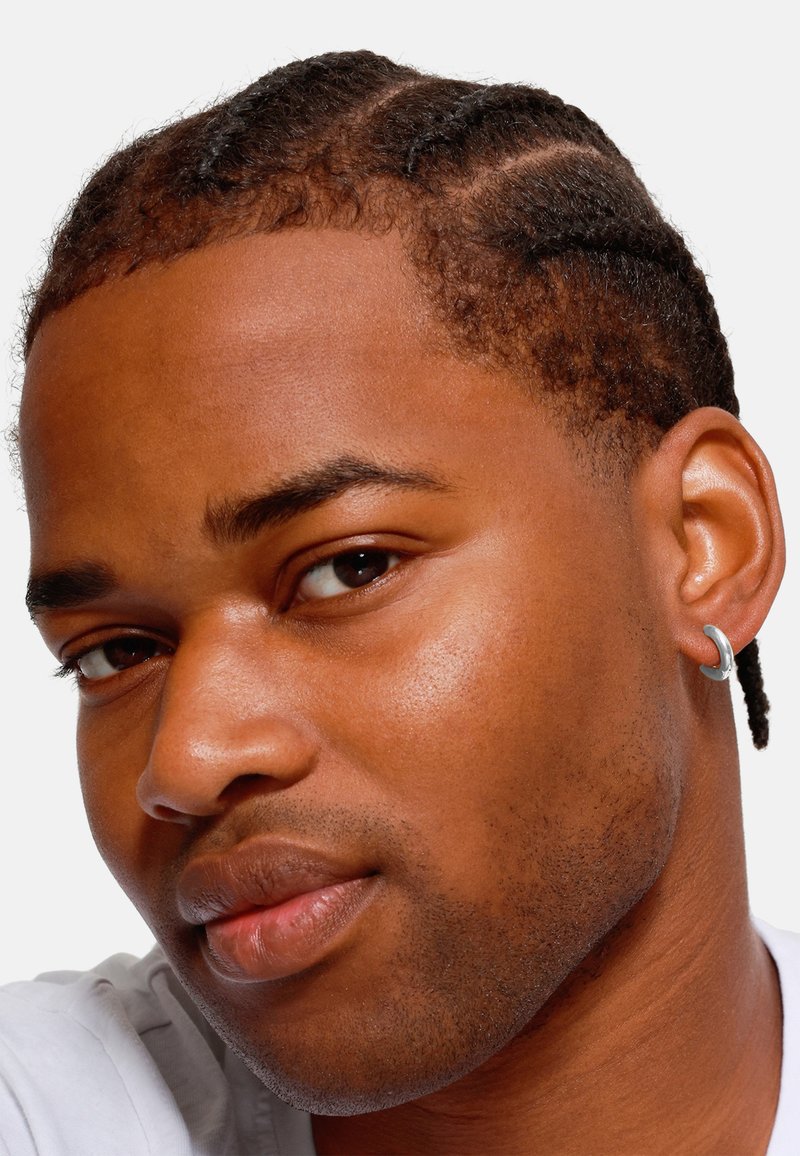 Gros plan d'un visage avec des cheveux tressés, une peau lisse et une petite boucle d'oreille en argent à l'oreille gauche, sur fond blanc uni.