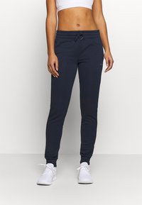 Mujer vistiendo pantalones deportivos azul marino con cintura ajustable, zapatos deportivos blancos y un top corto blanco, de pie contra un fondo liso.