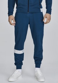 Blaue Sporthosen aus glattem Stoff, mit einem weißen Logo-Band am Knie, kombiniert mit weißen Turnschuhen und weißen Socken.