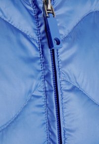 Giacca trapuntata azzurro chiaro con una texture liscia, una zip centrale con un gancetto blu e un motivo di cuciture a forma di diamante.
