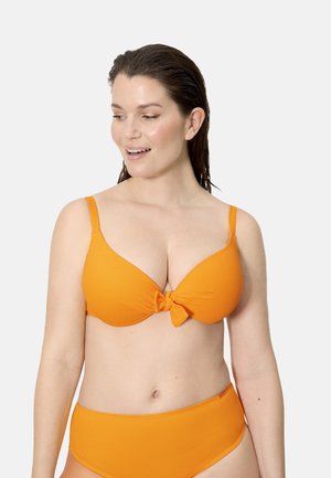 Mujer con el cabello mojado, vistiendo un bikini naranja con un detalle de nudo en la parte superior, mirando hacia abajo y sonriendo sobre un fondo liso.