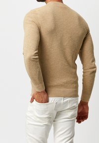 Beige stickad tröja med strukturerad yta, rund halsringning och långa ärmar, i kombination med vita byxor med bakfickor.