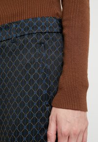 Manche de pull en maille côtelée marron associée à une jupe noire à motifs, ornée de formes en diamant bleues et dorées, texture lisse.