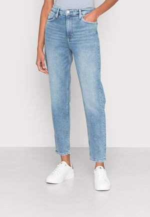 Person trägt hellblaue High-Waist-Jeans mit geradem Bein und weißen Sneakers, eine Hand in der Tasche, vor einem einfarbigen hellen Hintergrund.