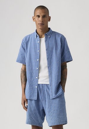 Mężczyzna z tatuażami ubrany w niebieską koszulę w kratę na krótkim rękawie zapinaną na guziki oraz pasujące krótkie spodenki, na białej prążkowanej koszulce na ramiączkach.