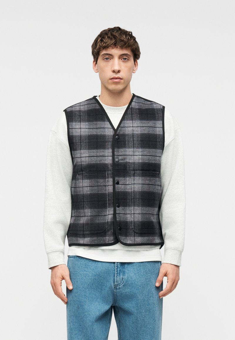 Νεαρός άνδρας που φοράει μαύρο και γκρι καρό γιλέκο με κουμπιά πάνω από λευκό φούτερ και μπλε τζιν, στέκεται μπροστά σε απλό φωτεινό φόντο.