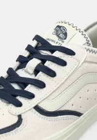 Λευκό sneaker από σουέτ με σκούρα μπλε κορδόνια και πλαϊνές λεπτομέρειες. Διαθέτει υφή στο επάνω μέρος, ανοιχτό γκρι λεπτομέρειες και ετικέτα με λογότυπο στη γλώσσα του παπουτσιού.