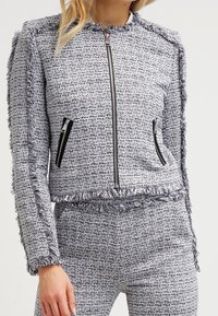 Grå och vit texturerad jacka med fransar, dragkedjefickor och utan krage. Materialet verkar vara vävt med ett subtilt mönster.