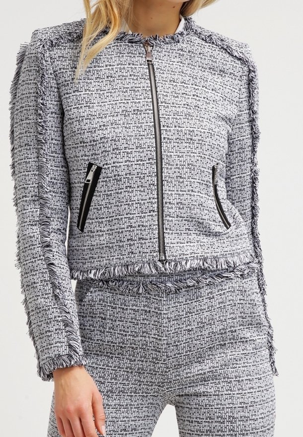 Grå och vit texturerad jacka med fransar, dragkedjefickor och utan krage. Materialet verkar vara vävt med ett subtilt mönster.