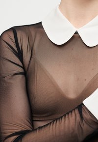 Άτομο που φοράει διάφανο μαύρο τοπ με λευκό γιακα τύπου Πίτερ Παν και αντίστοιχες μανσέτες, με σταυρωμένα τα χέρια στο ύψος του στήθους.