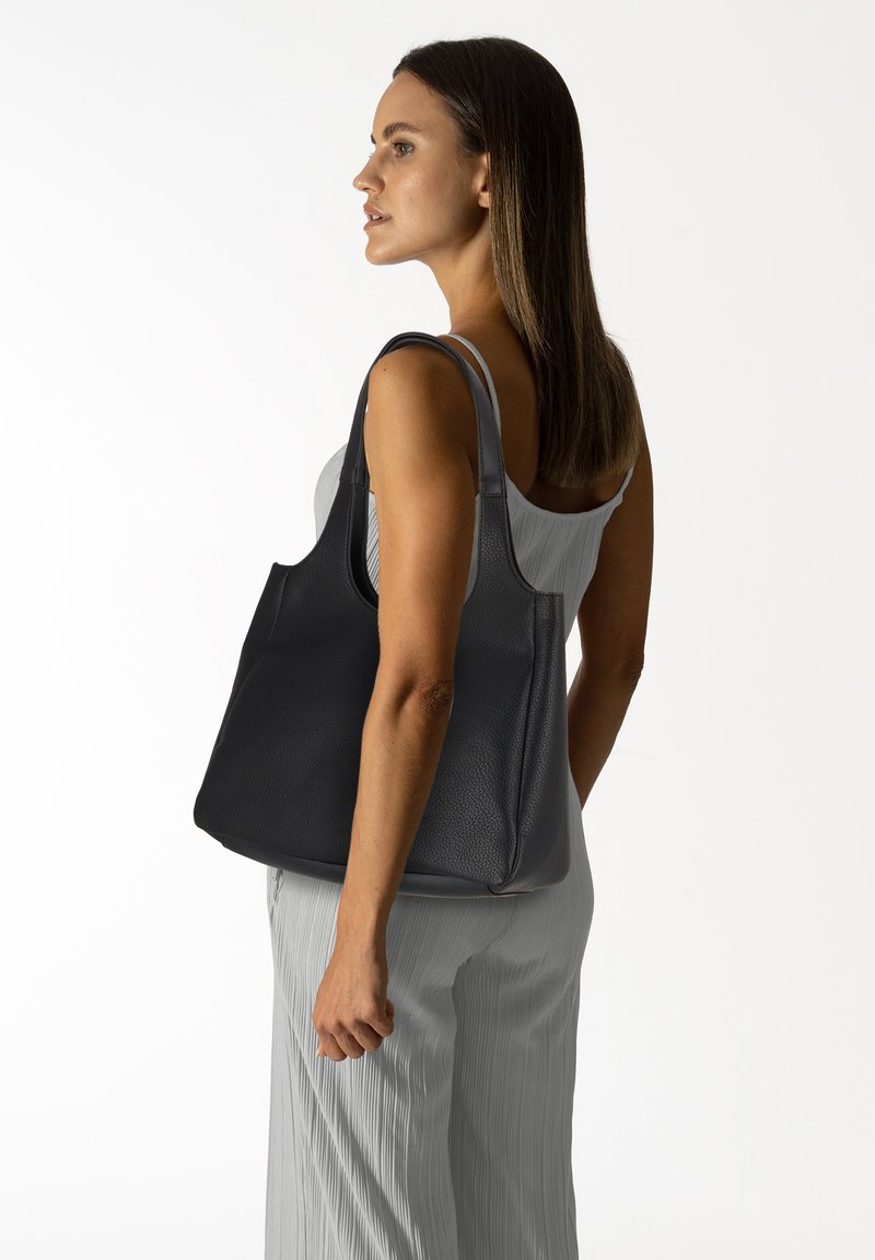 Sac de tote en cuir noir avec une surface texturée, des grandes poignées et une forme spacieuse, tenu par une personne portant des vêtements de couleur claire.
