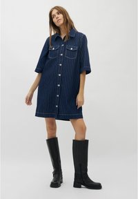 Jeanshemdkleid in Marineblau mit weißen vertikalen Streifen, kurzen Ärmeln, zwei Brusttaschen und silbernen Knöpfen; kombiniert mit kniehohen schwarzen Stiefeln.