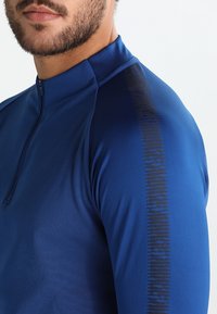 Maglietta sportiva blu a maniche lunghe con chiusura a mezza zip, texture liscia e una striscia grafica verticale nera lungo la spalla destra.