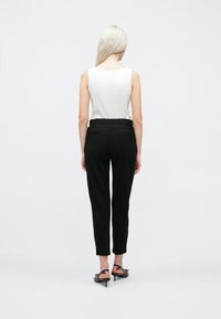 Crne tapered hlače s pojasom, uparene s bijelom topom bez rukava. Glatka tkanina i dizajn do gležnjeva. Vidljive visoke potpetice.