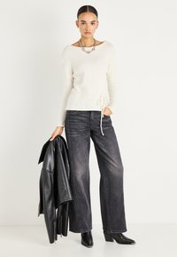 Femme portant un pull blanc, un jean noir large, des bottines noires, tenant une veste en cuir noire dans la main droite sur fond blanc.