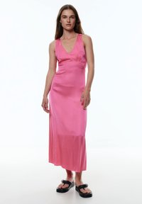 Rosa Satin-Maxi-Kleid mit V-Ausschnitt, tailliertem Oberteil und fließendem Rock; getragen mit schwarzen Plateau-Sandalen. Glatte Textur, minimalistisches Design.