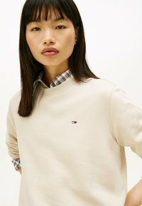 Felpa beige con scollo rotondo e maniche lunghe. Presenta un piccolo logo ricamato. Sotto, è visibile il colletto di una camicia a quadri.