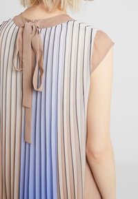 Femme portant une robe sans manches plissée avec des rayures verticales beige, blanches et bleues, nouée à l'arrière avec un nœud en ruban beige.