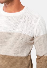 Pull en maille avec un motif texturé comportant trois bandes horizontales : blanche, crème et beige. Col rond, coupe décontractée.