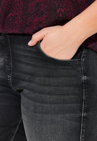 Main posé dans la poche avant d'un jean noir délavé, portant un haut à motifs bordeaux.