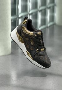 Czarny i złoty but sportowy z wzorzystą cholewką, metalicznymi akcentami, grubą białą podeszwą oraz kontrastującymi, teksturowanymi detalami po bokach.