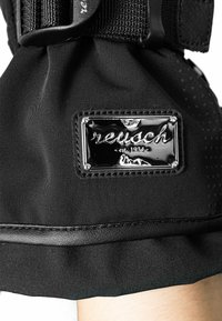 Zwarte stoffen handschoen met een glanzend zwart logo-patch met "reusch" en "est. 1934," aangevuld met gladde, getextureerde randen en stikdetails.