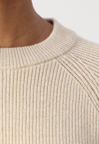 Μπεζ ριμπ πλεκτό πουλόβερ με στρογγυλή λαιμόκοψη και υφασμάτινη υφή, που αναδεικνύει μια μαλακή και ζεστή εμφάνιση με διακριτές ραφές.