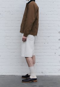 Giacca di tela marrone, pantaloncini di denim beige chiaro, scarpe scure con suole marroni e calzini bianchi. Profilo laterale contro un muro di mattoni bianco.