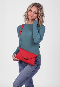 Taška ve stylu červené obálky vyrobená z texturované kůže se zlatým řetízkem, která má výrazný design sponky. Nosená přes tělo.