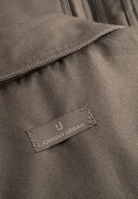 Hnedá tkanina s textúrovaným povrchom; obsahuje šitú etiketu so slovami "JOHNNY URBAN" a logom, čo prezentuje jednoduchý dizajn.